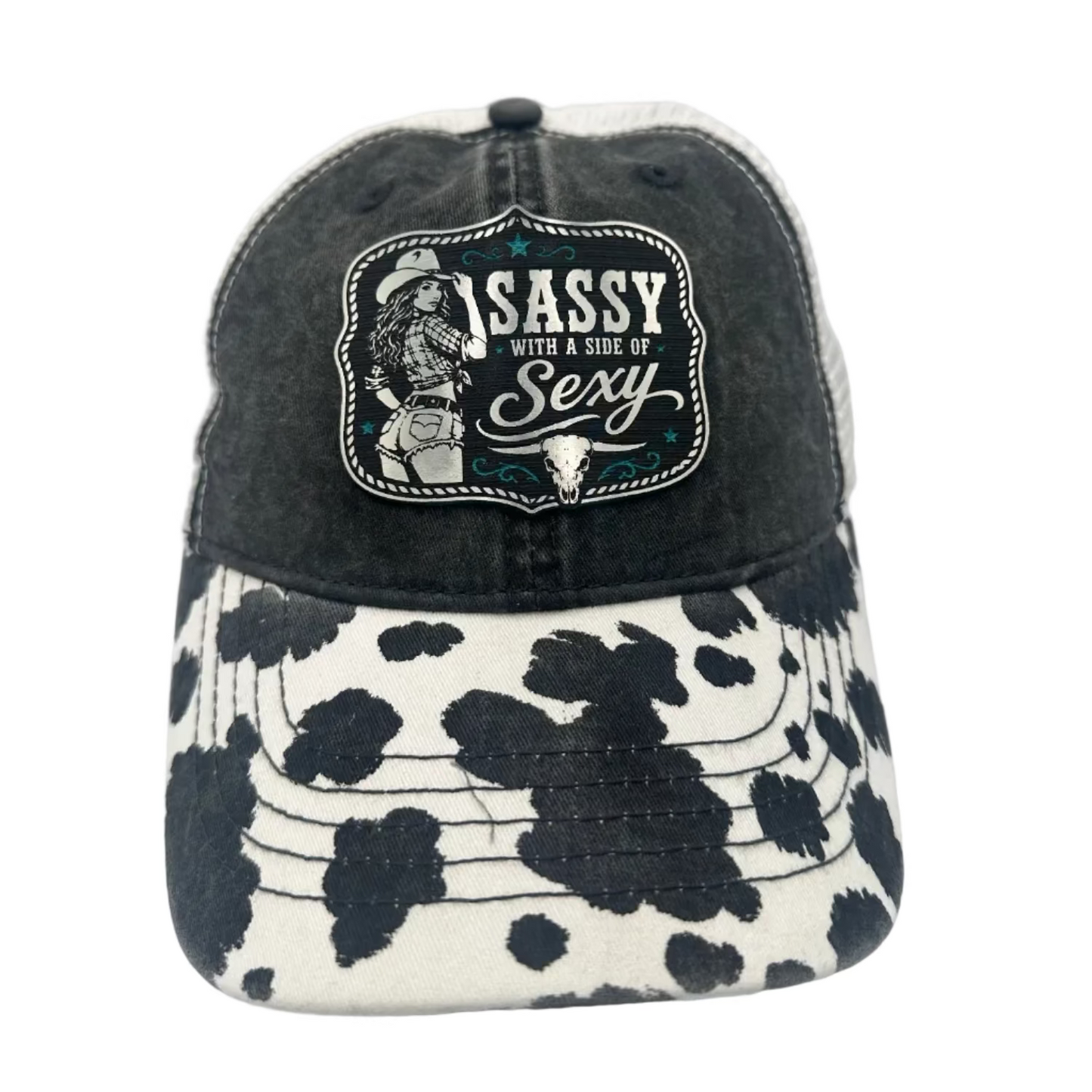 A distressed black trucker-style cap with a black-and-white cow-print brim and a front patch reading 'Sassy with a side of Sexy' with a cowboy illustration.