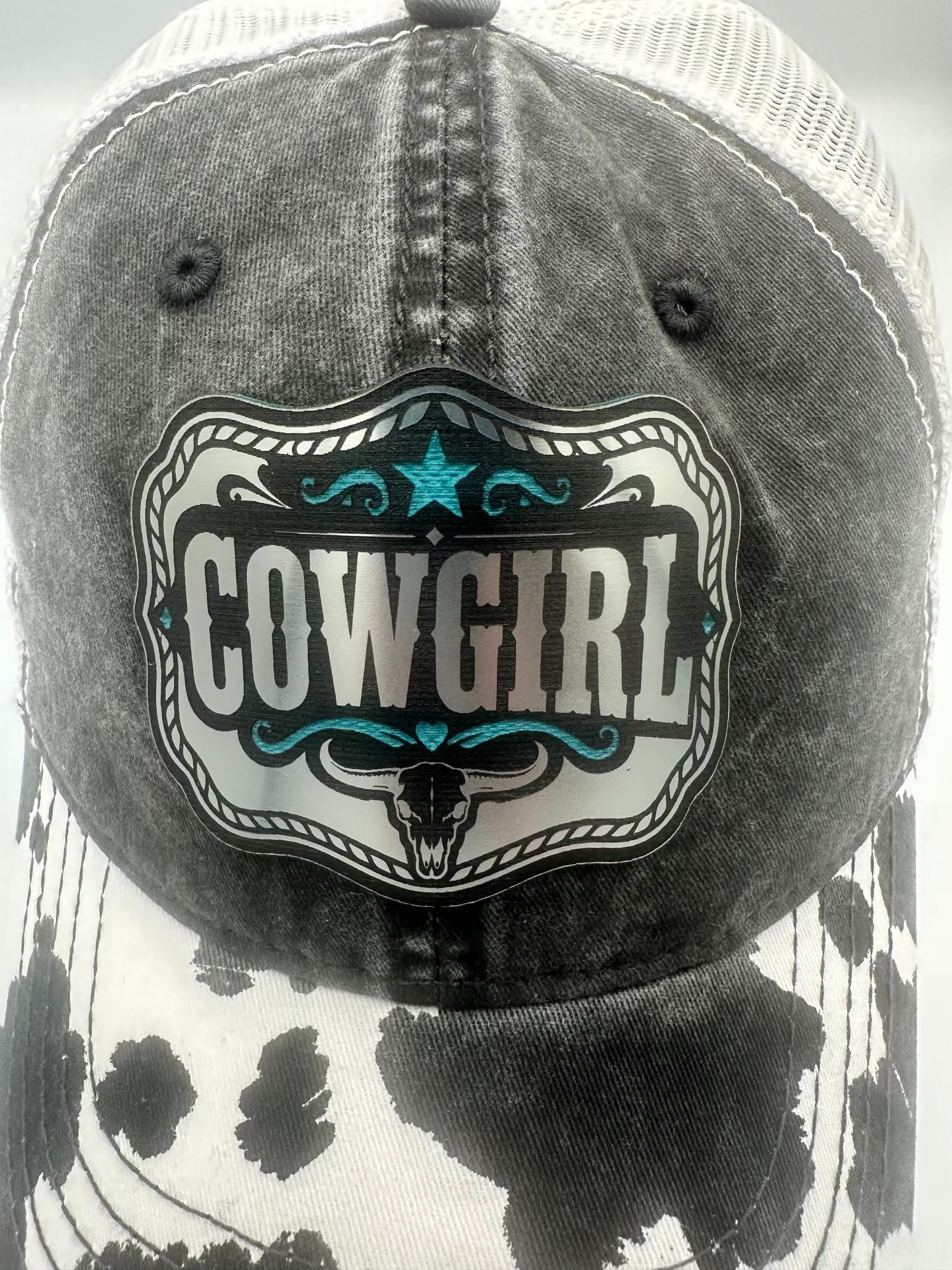 A close-up of a trucker-style baseball cap with a black-and-white cow-print front panel, white mesh back, and a large decorative front patch reading "COWGIRL" with turquoise accents.