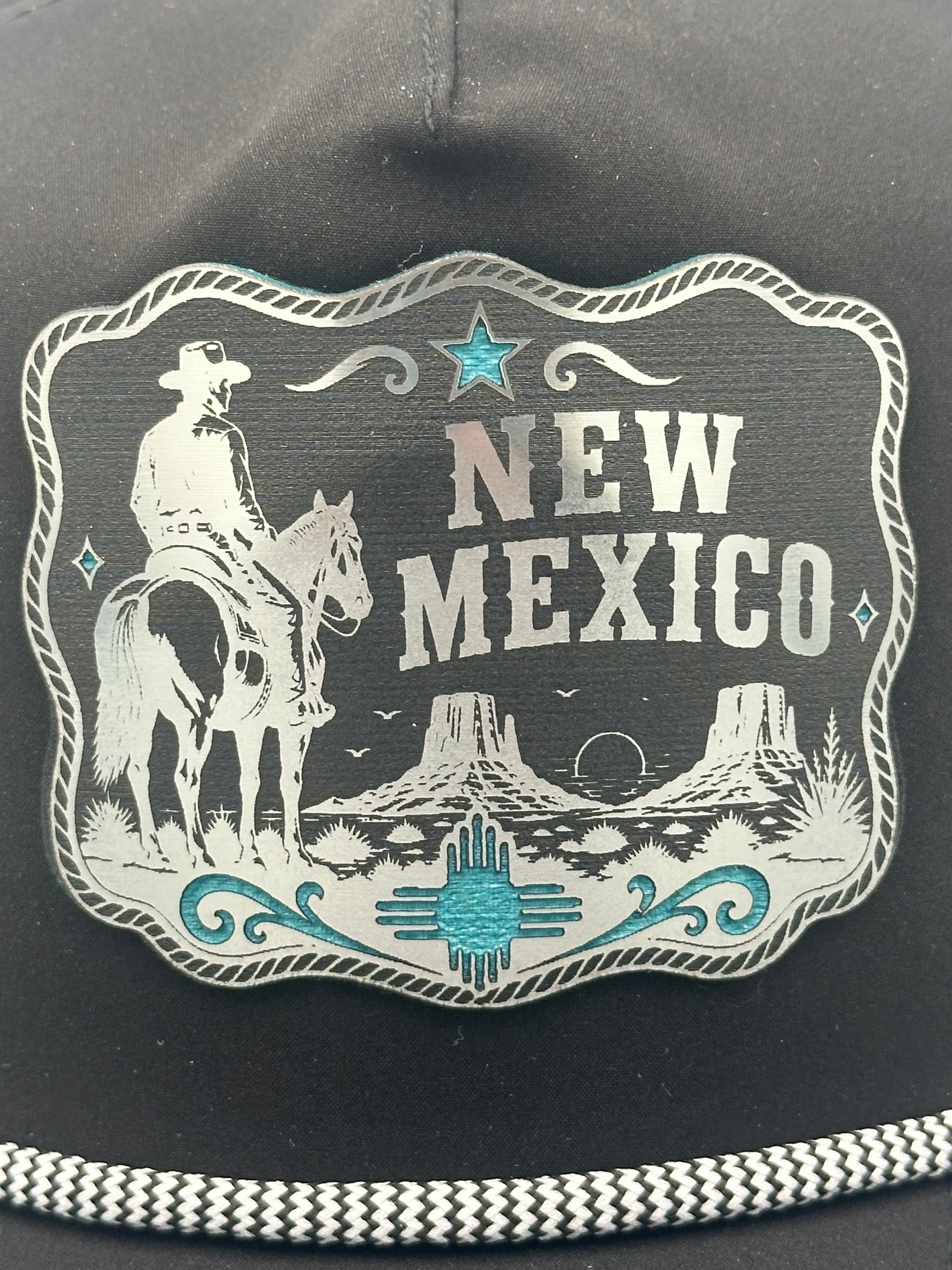 A close-up of a black hat featuring a large western-style metal patch with the words 'NEW MEXICO,' an illustration of a cowboy on horseback, desert mountains, and turquoise decorative accents. A braided black-and-white rope detail is visible along the lower edge.